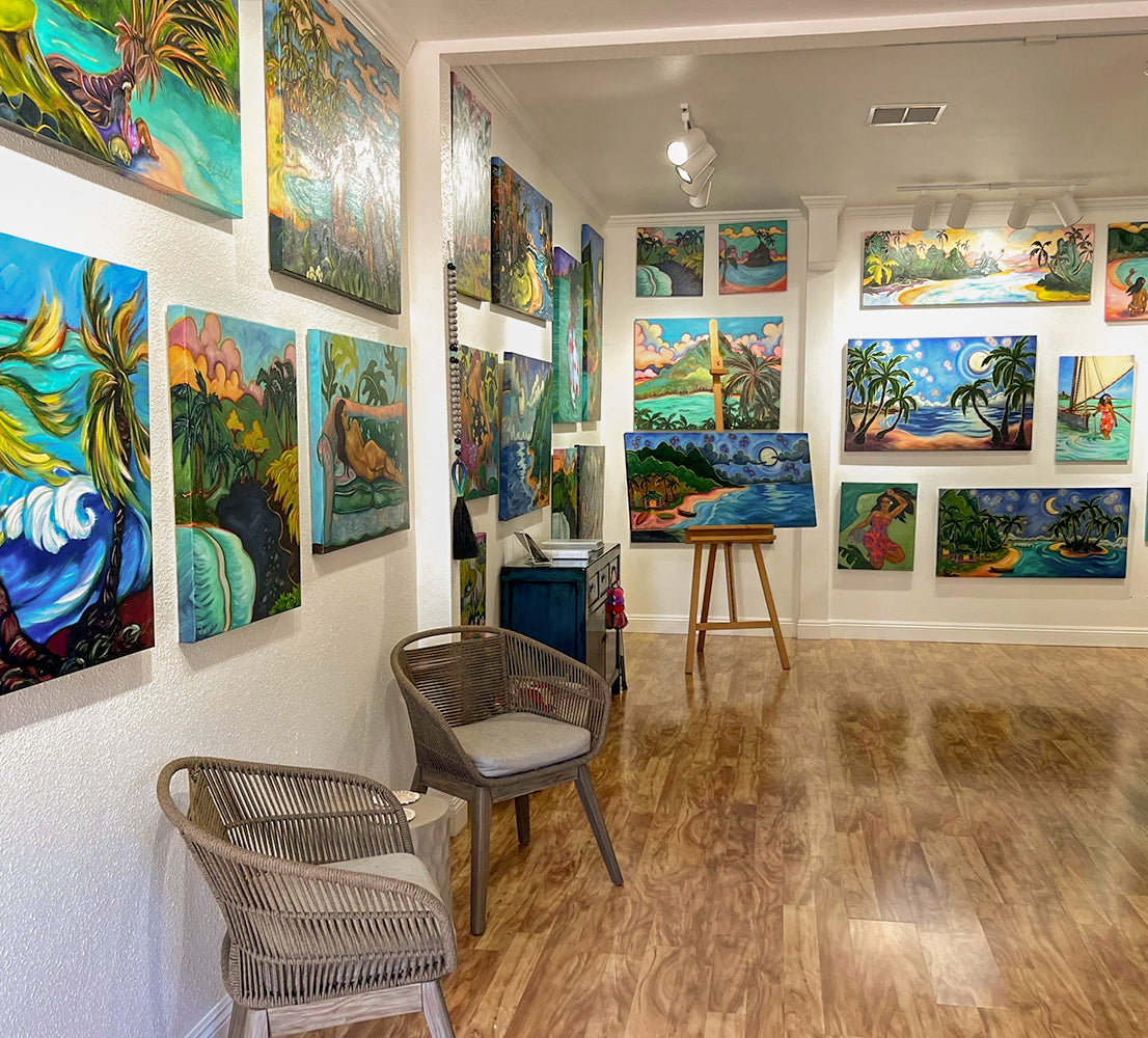 The Kim McDonald art gallery, a room filled with colorful tropical landscape paintings mounted on white walls, with two woven chairs in the foreground and a wooden stool near the center.