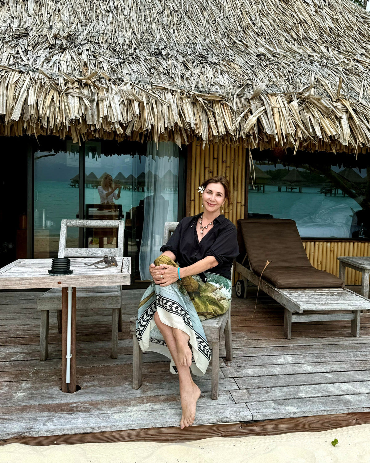 Kim sits barefoot on a wooden deck in front of a thatched-roof bungalow, holding a coconut. She wears a black short-sleeve top and a patterned skirt.