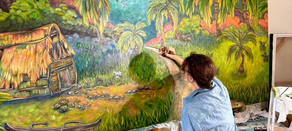 Kim McDonald sitting on a folding chair paints a large tropical landscape mural on a wall in an art studio. The painting depicts green mountains, palm trees, a thatched-roof hut, and a canoe under a blue sky.