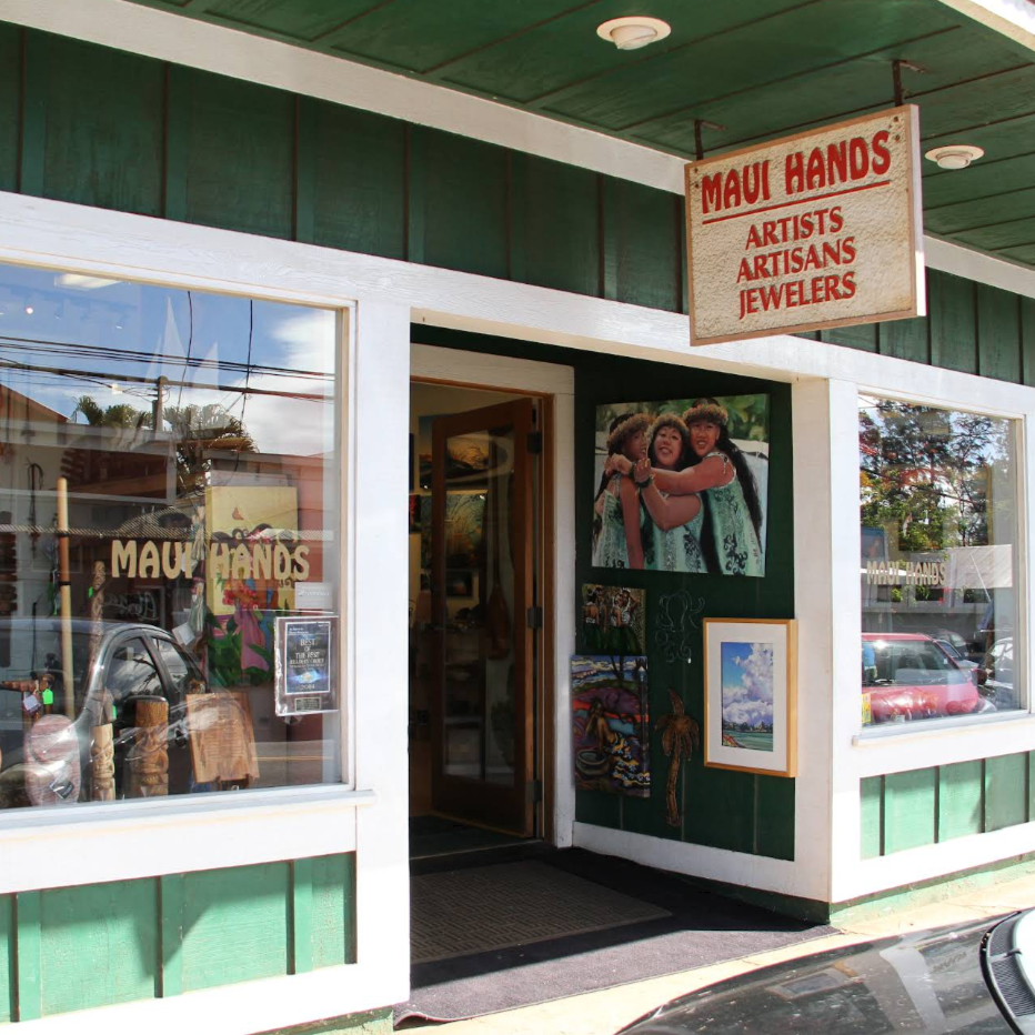Maui Hands Artists Artisans Jewelers storefront with a hanging wooden sign, green and white exterior, windows displaying jewelry, and a doorway in the center.