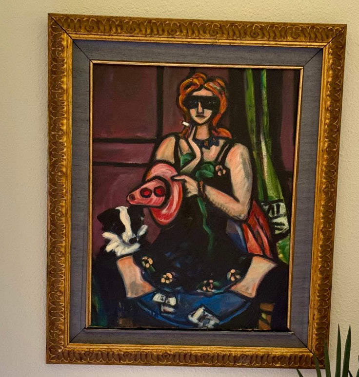 A framed painting hanging on a beige wall, depicting a stylized seated woman in a red headscarf and dark dress holding a pink object, with a gold ornate frame. A small woven-framed print hangs below, next to a potted plant and a window.